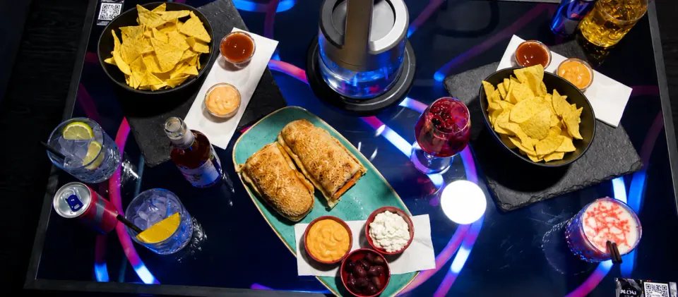 Derselbe Tisch gedeckt mit Nachos, überbackenem Sandwich, Dips und Drinks